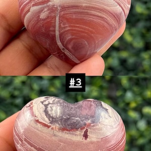 May include: Two heart-shaped stones with a swirling pattern of red, white, and purple hues. One stone has a sticker that reads "16 R", and the other has a sticker that reads "#3". The stones are held in a hand.