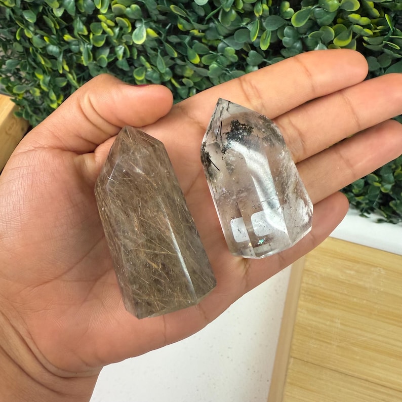 May include: Two quartz crystal towers, one brown with golden inclusions, the other clear with black inclusions. Both have pointed tops. The crystals are held in a hand.