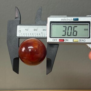 May include: A reddish-brown sphere is measured with a digital caliper. The digital display reads 30.6 mm. The caliper is grey and has a scale marked in millimetres and inches. The sphere appears to be polished.