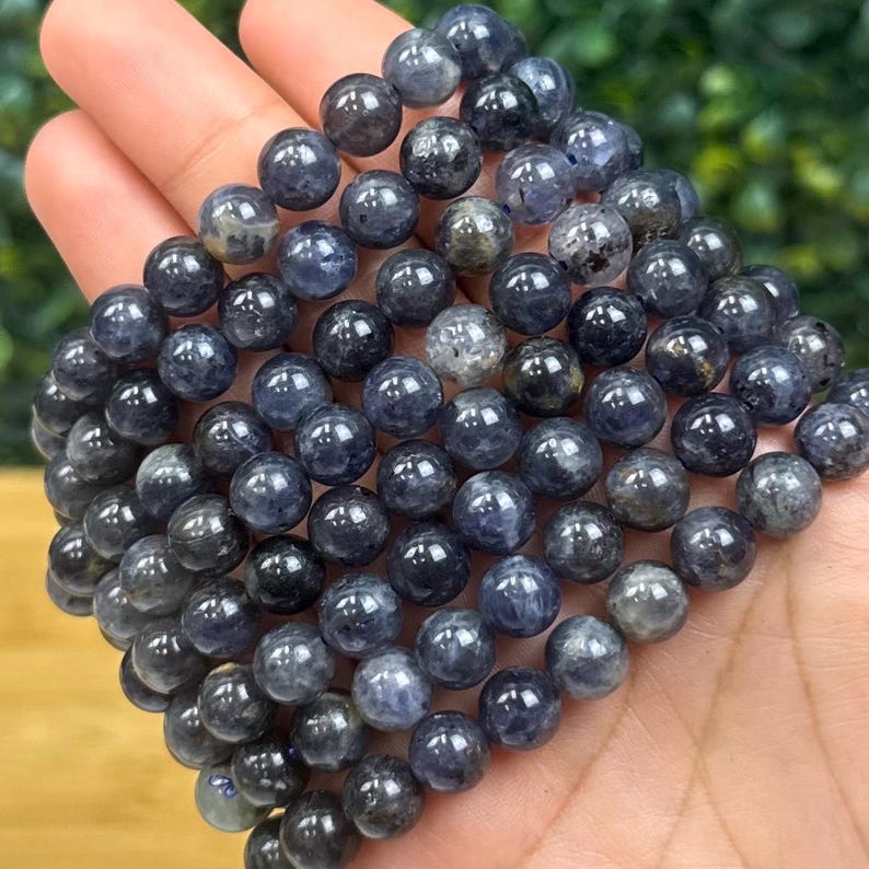 May include: A collection of dark blue, round gemstone beads. The beads are smooth and polished, with variations in color and texture. The beads are strung together, forming a bracelet or necklace. The gemstones are approximately 0.3 inches in diameter.