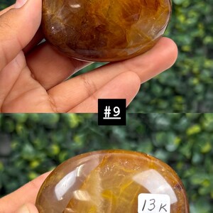 May include: A polished, oval-shaped gemstone with a warm, amber color and internal golden inclusions. The stone is held in a hand against a blurred green background. The text "#9" and "13K" are visible.