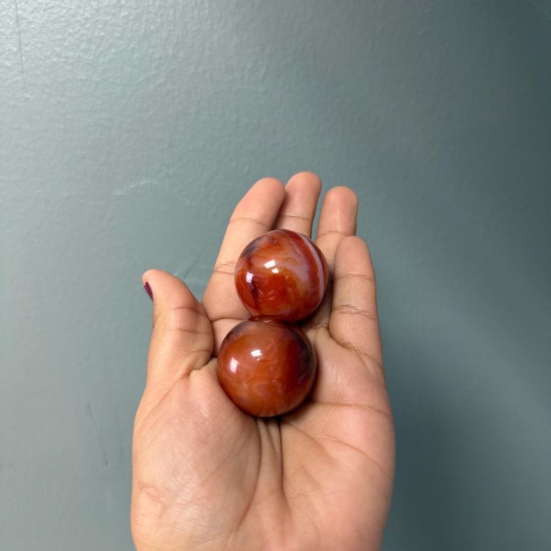 May include: Two polished, reddish-brown carnelian spheres sit in an open hand against a muted teal background. The spheres have a glossy finish and display natural patterns. The hand is in the foreground, with the background slightly out of focus.