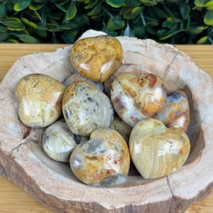 May include: A collection of heart-shaped stones in shades of tan, cream, and brown, with unique swirling patterns. The stones are displayed in a rustic wooden bowl, creating a natural and earthy aesthetic. The bowl sits on a light-colored wooden surface.