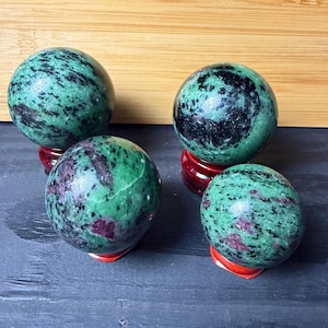 Könnte beinhalten: Vier polierte Edelstein-Kugeln. Die Kugeln sind hauptsächlich grün mit schwarzen und roten Einschlüssen. Jede Kugel ruht auf einem kleinen, roten, dekorativen Ständer. Der Hintergrund zeigt eine Holzoberfläche und eine dunkelgraue Oberfläche.