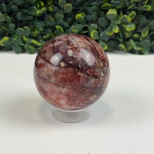 May include: A polished, spherical gemstone with a mix of red, brown, and clear hues. The crystal ball sits on a clear, round stand. The background features green foliage.