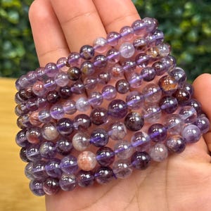 May include: A stack of amethyst gemstone bracelets. The bracelets are made of round, polished beads in shades of purple, lavender, and brown. The beads are strung together on a stretchy cord. The bracelets are held in a hand.