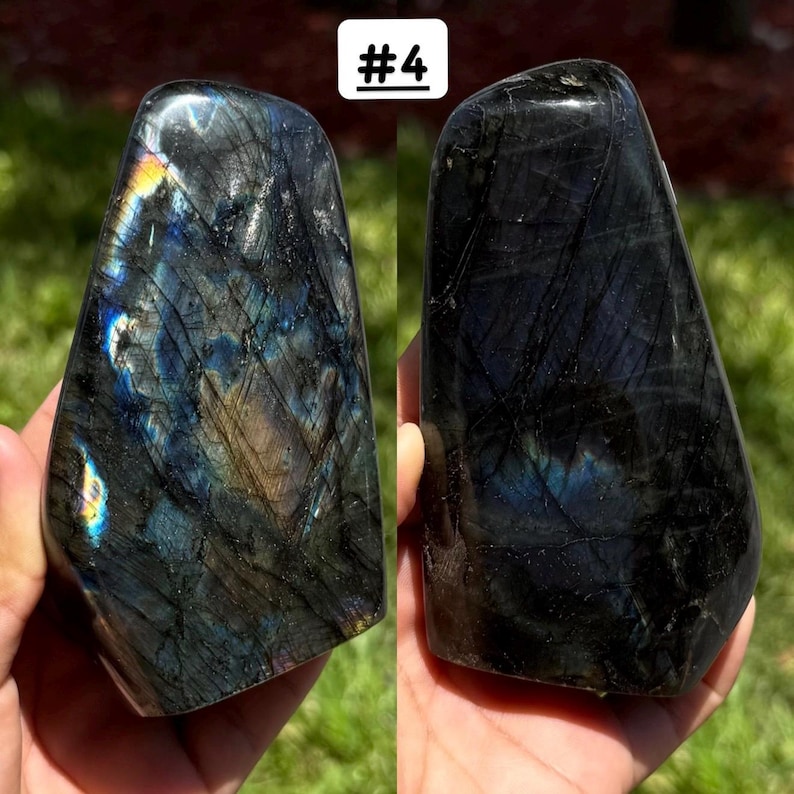 May include: Two polished labradorite stones labeled #4, featuring dark gray and black surfaces with iridescent flashes of blue, gold, and yellow light reflecting from within the crystalline structure, showcasing natural fissures and smooth, rounded edges.