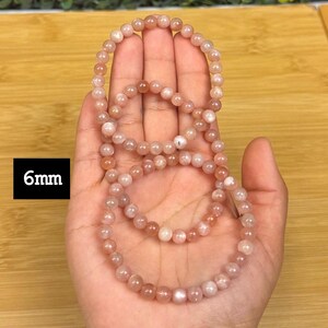 Op de afbeelding: Drie strengen van 6 mm zonnesteen kralenarmbanden worden in een hand getoond. De kralen hebben een licht perzikkleur met een subtiele glans. De armbanden liggen op een houten oppervlak.