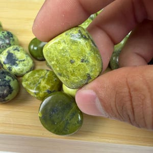 May include: A hand holding a smooth, rounded, lime green stone with dark green and black markings. Other similar stones are scattered on a wooden surface. The stones have a polished appearance.