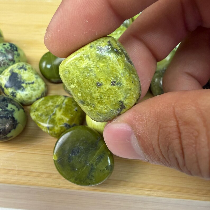 May include: A hand holding a smooth, rounded, lime green stone with dark green and black markings. Other similar stones are scattered on a wooden surface. The stones have a polished appearance.