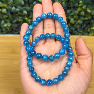 May include: Two blue apatite beaded bracelets. The bracelets are made of round, polished beads in varying shades of blue. The bracelets are displayed on a hand, with a blurred green background.