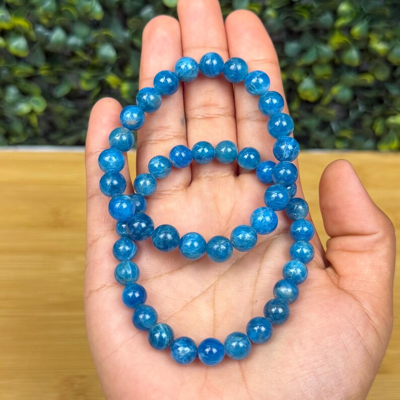 May include: Two blue apatite beaded bracelets. The bracelets are made of round, polished beads in varying shades of blue. The bracelets are displayed on a hand, with a blurred green background.