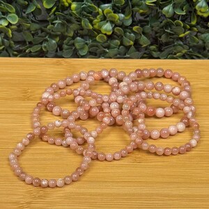 Op de afbeelding: Verschillende armbanden gemaakt van kleine, ronde, roze-oranje kralen. De armbanden zijn in een cirkelvorm op een houten oppervlak gerangschikt. De kralen hebben een gladde textuur en een subtiele glans.