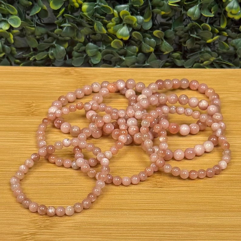 Op de afbeelding: Verschillende armbanden gemaakt van kleine, ronde, roze-oranje kralen. De armbanden zijn in een cirkelvorm op een houten oppervlak gerangschikt. De kralen hebben een gladde textuur en een subtiele glans.