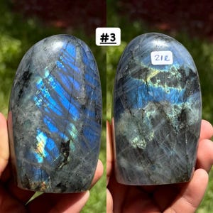 May include: A polished labradorite stone with a smooth, rounded top and a flat base. The surface displays dark gray tones with vibrant blue and yellow iridescent flashes. The stone on the right features a white label with the text 21R.