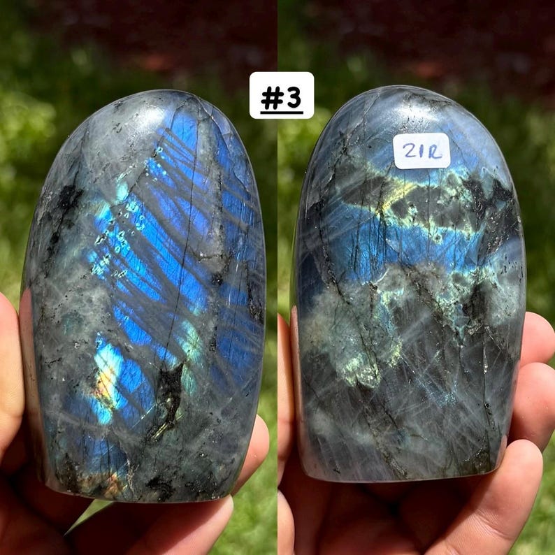 May include: A polished labradorite stone with a smooth, rounded top and a flat base. The surface displays dark gray tones with vibrant blue and yellow iridescent flashes. The stone on the right features a white label with the text 21R.