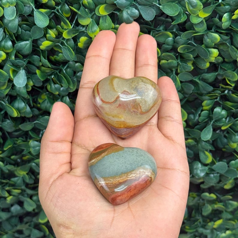 Puede incluir: Dos piedras en forma de coraz&oacute;n con patrones naturales en tonos marr&oacute;n, beige, rojo y verde. Las piedras se sostienen en una mano abierta sobre un fondo de follaje verde. Las piedras pulidas son probablemente decorativas o para exhibici&oacute;n.