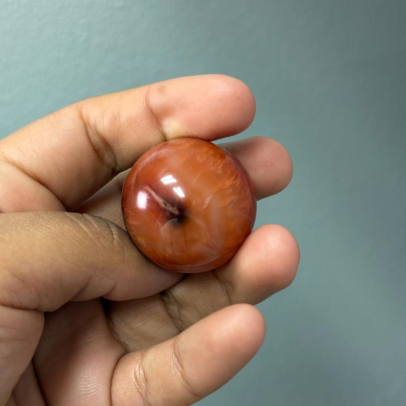May include: A polished, spherical gemstone in a warm, reddish-orange colour. The stone has a smooth, glossy surface with subtle internal patterns and a small, dark inclusion. The gemstone is held in a hand against a muted blue-green background.