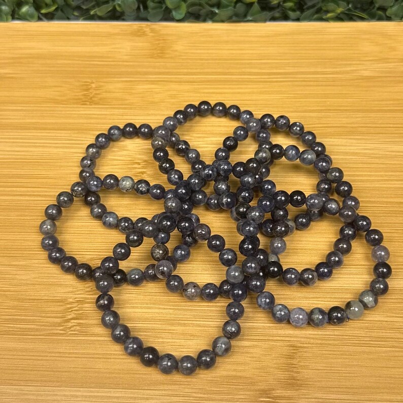 May include: Several dark blue beaded bracelets are arranged on a wooden surface. Each bracelet is made of small, round beads. The bracelets are a mix of dark blue and gray tones, creating a subtle color variation.