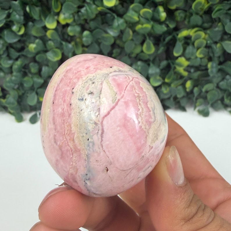 May include: A polished, egg-shaped gemstone with a pink and cream color scheme. The stone has a marbled appearance with visible veins and variations in color. The surface is smooth and reflective, suggesting a high level of polish.