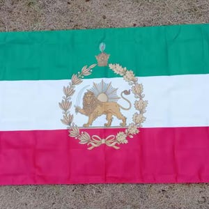 Könnte beinhalten: Rechteckige Flagge mit horizontalen Streifen in Grün, Weiß und Rot. In der Mitte befindet sich ein detailliertes Emblem mit einem Löwen, einem Schwert und einer Sonne, umgeben von einem Kranz und einer Krone. Die Flagge ist aus Stoff.