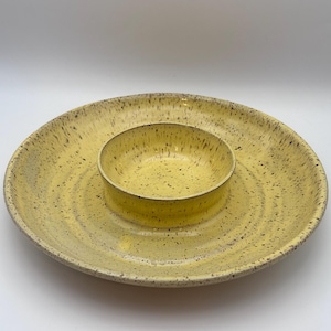 May include: A yellow ceramic serving dish with a central bowl. The dish has a speckled pattern of dark brown spots. The outer rim is wide and flat, while the inner bowl is smaller and circular. The dish is designed for serving snacks or dips.
