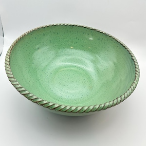 Può includere: Una ciotola verde in ceramica con un interno maculato e un motivo a corda sul bordo. La ciotola ha una forma ampia e poco profonda e una finitura lucida. Il colore è un verde tenue e smorzato.