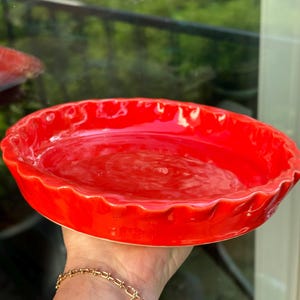 Puede incluir: Un plato de tarta de cerámica rojo brillante con un borde festoneado. El plato es redondo y tiene un acabado brillante. El plato de tarta se sostiene frente a una ventana con un fondo verde.
