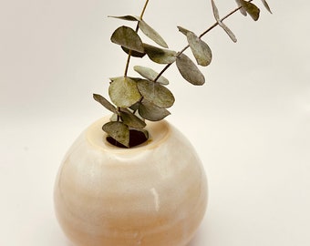 May include: A small, round, off-white ceramic vase with a wide opening, holding a sprig of dried eucalyptus.