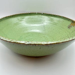 May include: A handmade ceramic bowl with a light green glaze. The bowl has a wide, shallow shape with a textured rim and a speckled interior. The exterior is a darker green, and the bowl appears to be made of stoneware.