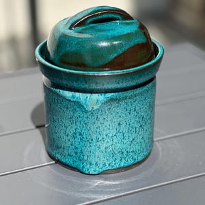 Puede incluir: Una mantequera de cerámica artesanal con esmalte turquesa y marrón. El plato tiene una base redonda y una tapa a juego con asa. El exterior presenta un acabado texturizado y moteado. La mantequera está diseñada para contener un tamaño de mantequilla estándar.