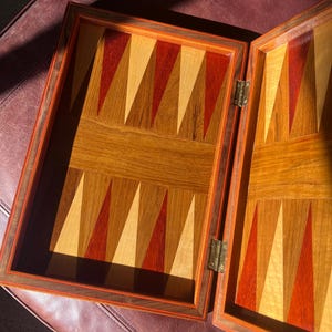 May include: An open wooden backgammon board featuring a geometric inlay pattern of alternating red and light tan triangular points set against a natural wood grain background, connected by two brass hinges on the side.