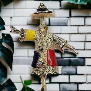 May include: A gold-toned scarecrow brooch with a yellow, red, and blue patterned coat and a black hat.