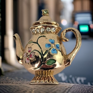 May include: A gold teapot with a floral design. The teapot has blue, pink, and green flowers on it. The teapot is decorated with rhinestones.
