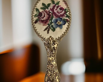 Puede incluir: Un espejo de mano vintage con un mango dorado y un marco ovalado blanco con un diseño floral bordado con flores rosas, moradas y azules.