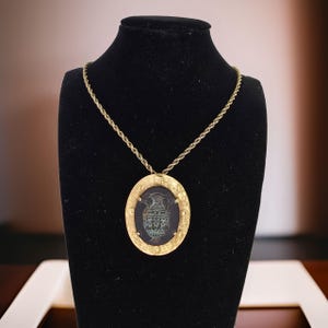 May include: Gold-toned pendant necklace featuring an oval-shaped pendant with a dark background and a detailed, teal-colored vase design. The pendant is framed by a decorative gold border and hangs from a matching gold rope chain. The necklace is displayed on a black mannequin.