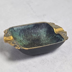 May include: A small, rectangular ashtray with a dark interior and ornate gold-colored edges. The interior has a green patina, and the ashtray has a small rest for a cigarette. The ashtray is made of metal.