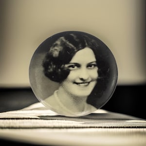 Puede incluir: Una fotografía en blanco y negro de una mujer con el pelo corto y ondulado. Ella sonríe y lleva una blusa blanca. La fotografía está en un marco redondo y transparente.