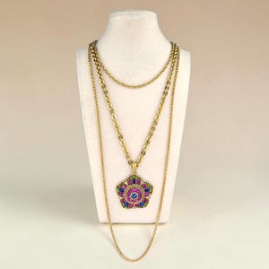 Könnte beinhalten: Eine mehrsträngige goldfarbene Halskette mit einem großen, fünfblättrigen Blumenanhänger. Der Anhänger ist mit leuchtend pinken, blauen und grünen Strasssteinen besetzt. Die Kette besteht aus drei verschiedenen Goldketten unterschiedlicher Länge.