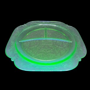 May include: A vintage, divided serving dish in a vibrant, glowing green hue. The glass dish features an oval center section with a divider and ornate, embossed floral patterns. The edges have a scalloped design.