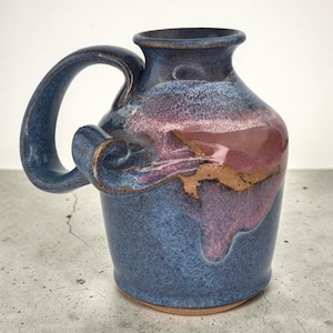 May include: A ceramic mug featuring a wide, curved handle and a flared rim. The exterior is finished with a mottled blue glaze, accented by a prominent patch of deep pink and a streak of earthy brown running across the center of the vessel body.