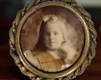 May include: Vintage, circular, brass-coloured picture frame featuring a sepia-toned portrait of a young girl. The frame has a decorative, rope-like edge. The girl wears a bow in her hair and a light-coloured dress.
