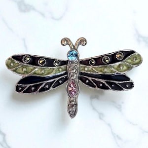 May include: A silver-toned dragonfly brooch with black, green, and purple enamel wings. The brooch features a light blue gemstone at the head, a pink gemstone on the body, and small, round, silver-colored accents. The wings have a layered design.