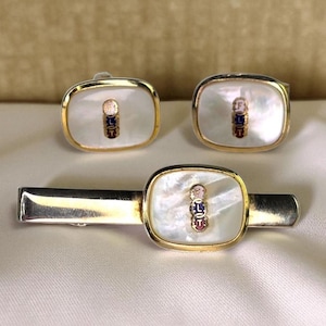 May include: A vintage jewelry set featuring a tie clip and a pair of cufflinks. Each piece has a gold-tone frame with a mother-of-pearl inlay. The inlay features a small, colorful emblem. The set is displayed on a white surface.