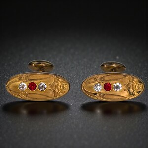 May include: A pair of antique gold cufflinks with an oval design. Each cufflink features three gemstones: a central red stone flanked by two clear stones. The gold setting has an ornate floral pattern.