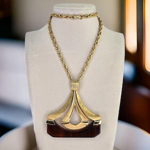 May include: A gold necklace with a statement pendant. The pendant has a geometric design with a brown rectangular base and gold detailing. The necklace is displayed on a white mannequin.