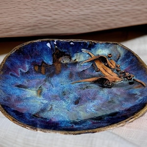 Op de afbeelding: Een klein, ovaal keramisch schaaltje met een blauwe en paarse glazuur. Het schaaltje heeft een reliëfdesign van een eucalyptus tak met bladeren en bessen.