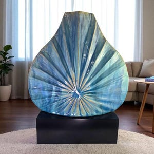 May include: Large, abstract art piece in shades of blue and gold, resembling a stylized leaf or fan. The artwork is displayed on a black rectangular base, creating a striking contrast. The piece is a decorative element for a living space.