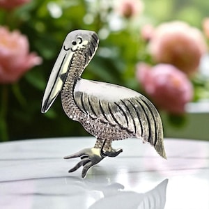 Pode incluir: Broche de pelicano prateado. A ave é retratada com penas detalhadas e um longo bico. O broche está sobre uma superfície com um fundo desfocado de flores cor-de-rosa e vegetação.