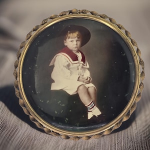 May include: A vintage framed photo of a young boy wearing a sailor outfit and a hat. The photo is in a gold-colored frame with a rope-like design.